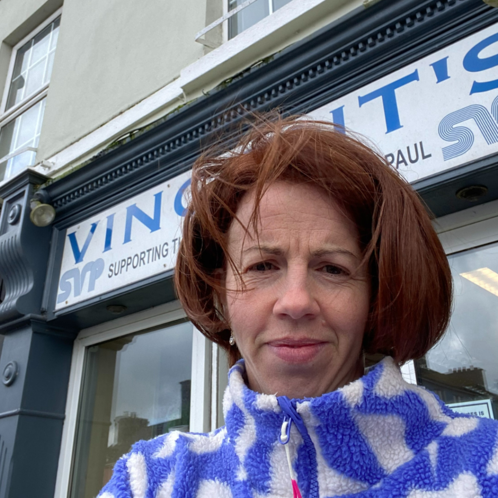 Anne Marie Kingston of White Sage Decluttering, a lady with red hair is taking a selfie in front of a St. Vincent de Paul charity shop. The sign above reads "Vincent's SVP Supporting." Anne Marie is wearing a blue and white patterned jacket, and the overcast sky looms above, adding a cozy charm to their shot.