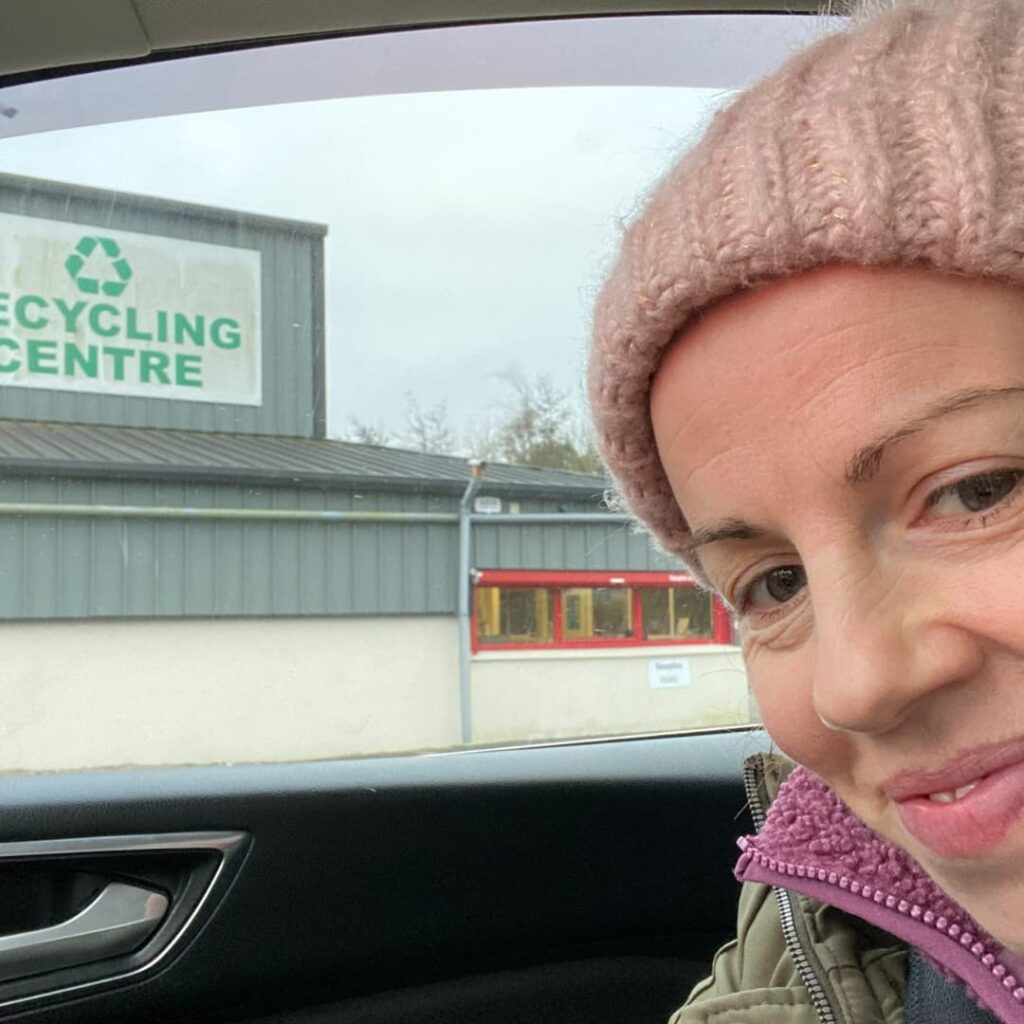 Anne Marie Kingston founder of White Sage Decluttering is wearing a pink knit hat and jacket is smiling inside a car parked outside the Recycling Centre. The building, with its welcoming sign, seems like the perfect place to drop off your clutter after following Anne Marie's attic decluttering tips.