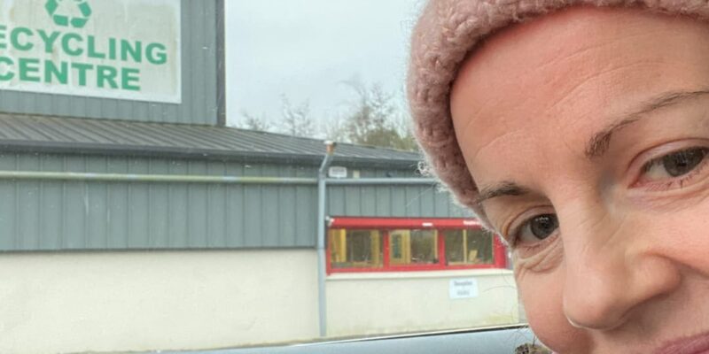 Anne Marie Kingston wearing a pink knit hat and jacket is smiling inside a car parked outside the Recycling Centre. The building, with its welcoming sign, seems like the perfect place to start after her tackling attic decluttering tips.