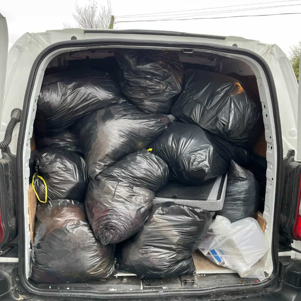 A van with its back doors open, revealing it fully loaded with numerous black garbage bags. The bags are tightly packed, filling the space completely. A few small items are wedged in the gaps. Overcast sky in the background.