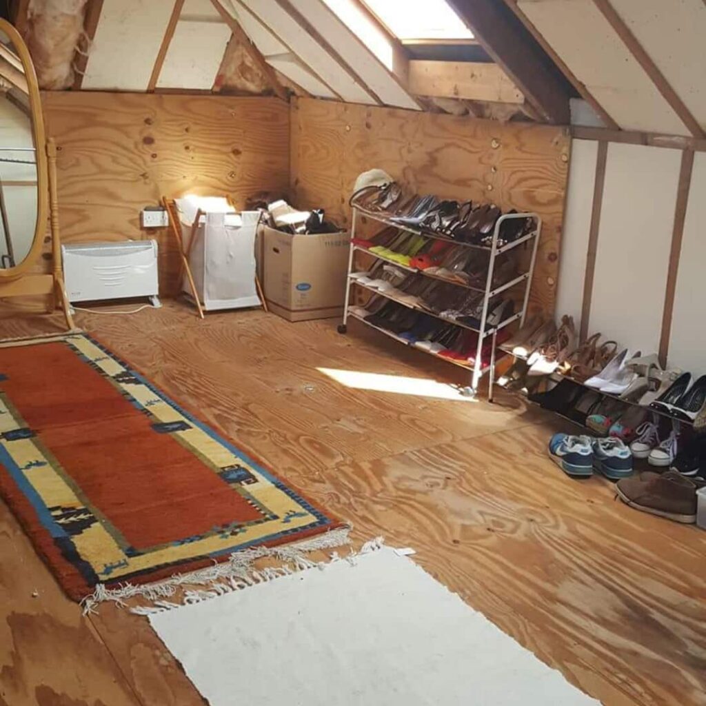 A cozy attic space with wooden walls and floor features a colourful rug and white mat. A shoe rack filled with various shoes leans against the wall. This attic has been decluttered by Anne Marie Kingston of White Sage Decluttering following her attic decluttering tips. 